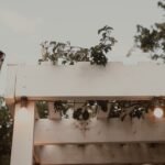 Backyard wedding ideas with a pergola strung with warm string lights and an outdoor umbrella