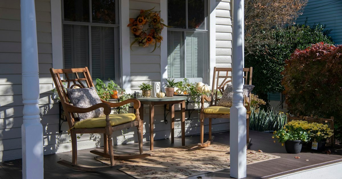 Fall porch decor with wooden chairs, pumpkins, and mums near a white porch post