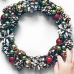 Dollar Tree DIY Christmas decorations: hands building a wreath from pinecones and red and gold baubles on a table