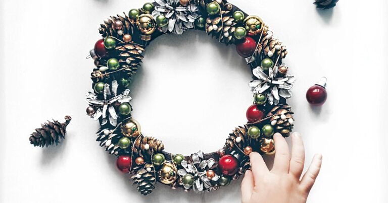 Dollar Tree DIY Christmas decorations: hands building a wreath from pinecones and red and gold baubles on a table