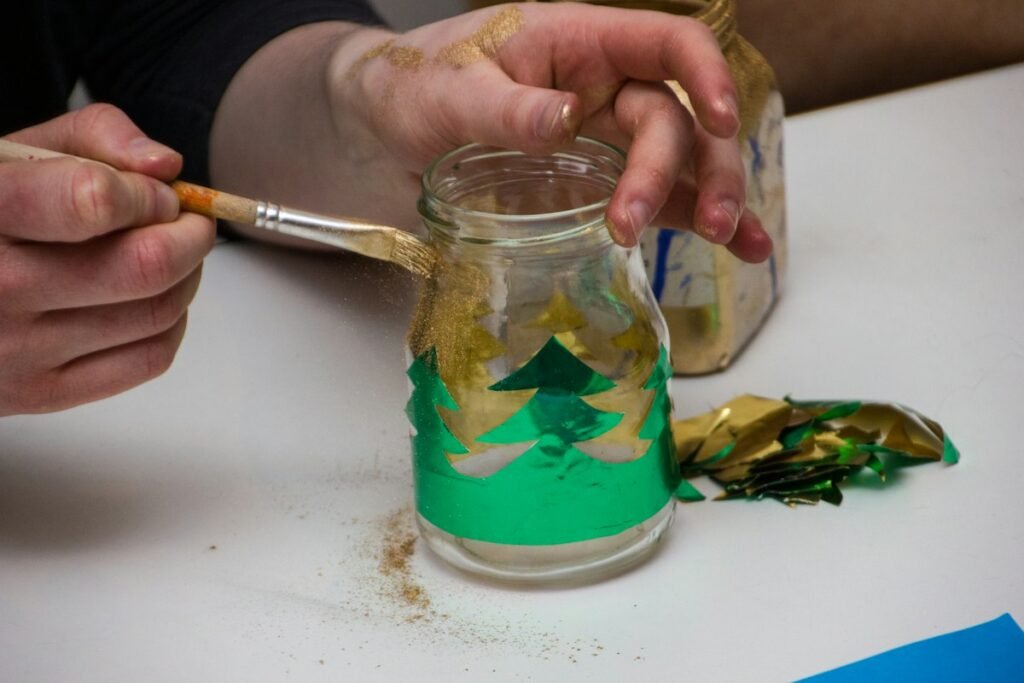 Hands painting a glass jar green and gold, one of the easiest dollar tree diy christmas decorations