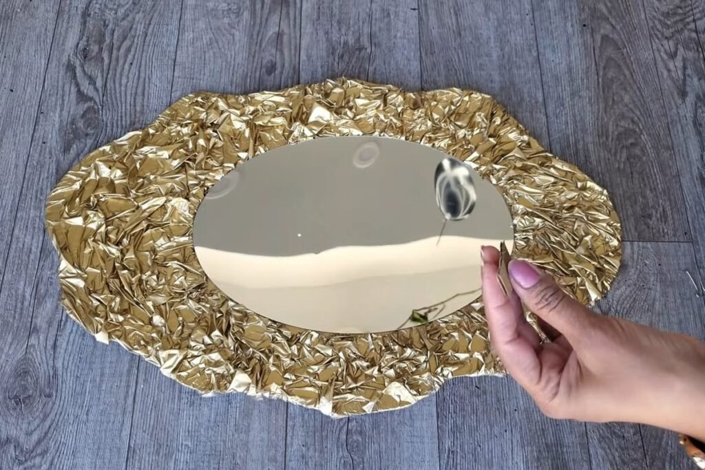 A hand holding a small binder clip next to a finished oval mirror in a crinkled gold frame