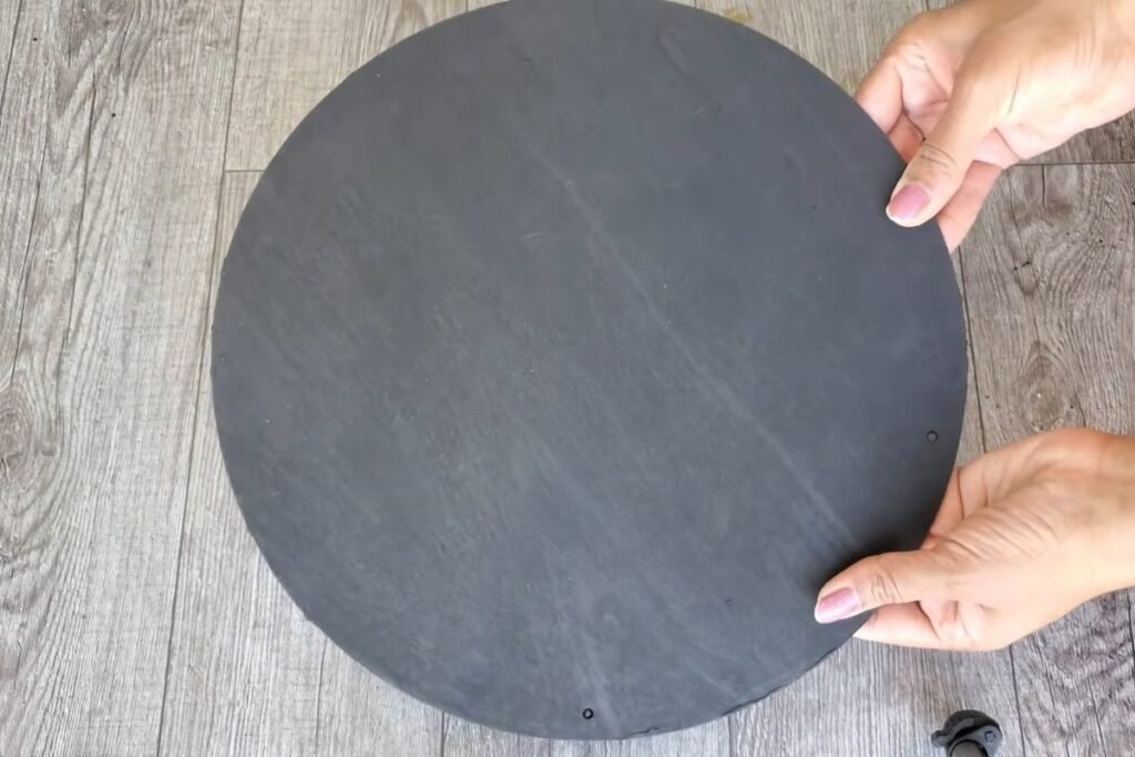 A hand holding a round wooden board that has been painted flat black on a grey wood floor