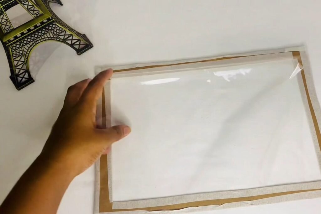 Hand smoothing a sheet of clear plastic over a tape-wrapped cardboard frame