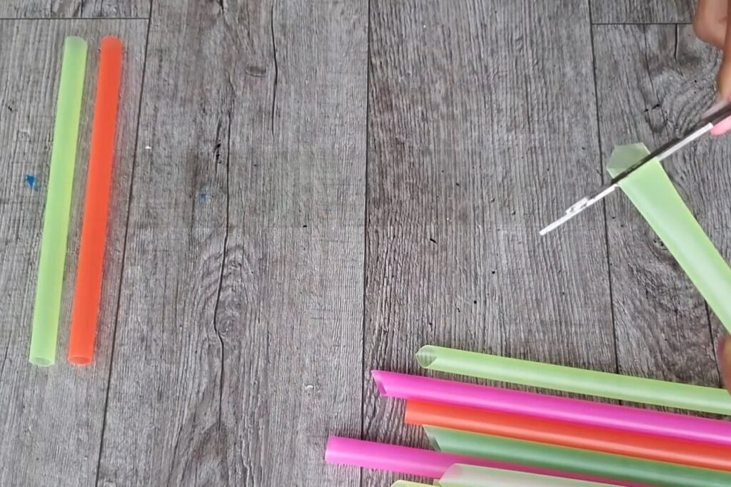 A hand holding scissors cutting a green jumbo straw beside a pile of colored straws