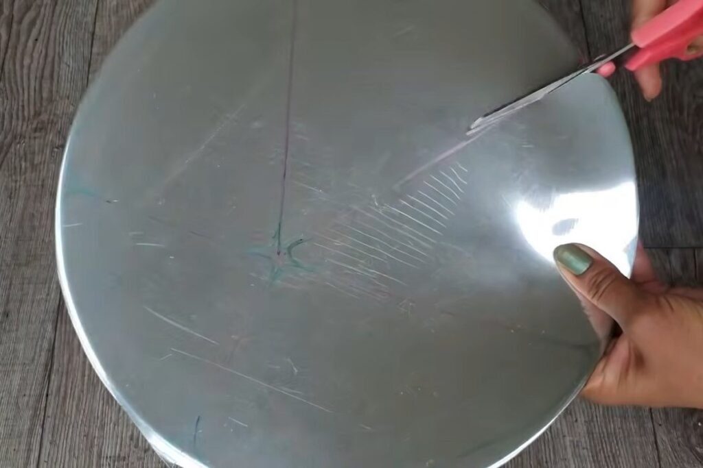Pink scissors cutting along a marked line through a circular metal tray held in one hand