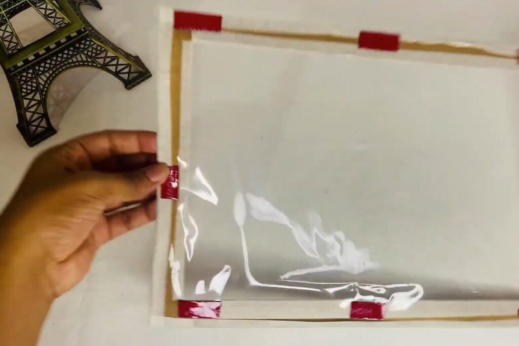Hand lifting clear plastic to show short red double-sided tape pieces on a frame ring