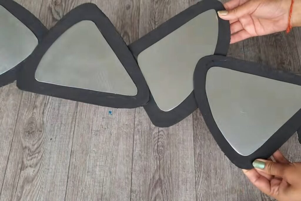 Four connected black-framed rounded triangle mirrors laid out on a wooden floor with a hand holding the edge