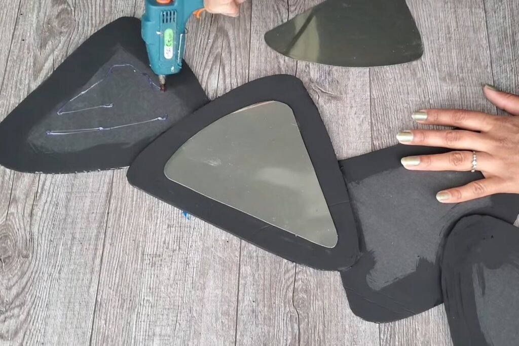 A hot glue gun and a hand pressing a rounded triangle mirror into a black foam frame in a connected chain