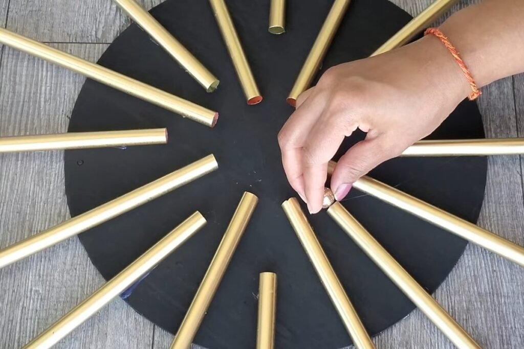 A black circle covered with many gold straws radiating outward while a hand places one more straw