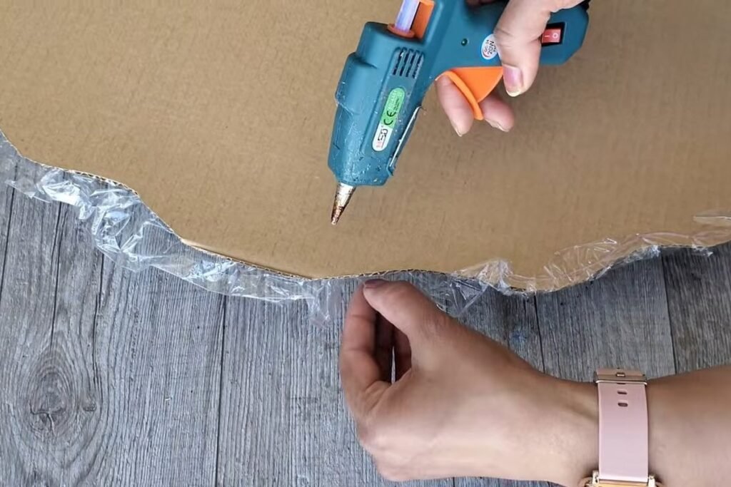 The back of a cardboard piece with a glue gun being used to secure plastic wrap that wraps over the edge