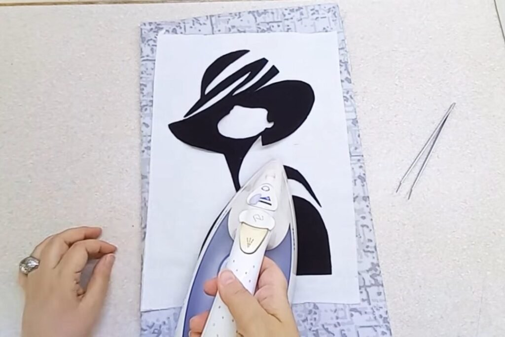 An iron pressing black fabric shapes onto a white panel on a patterned ironing surface