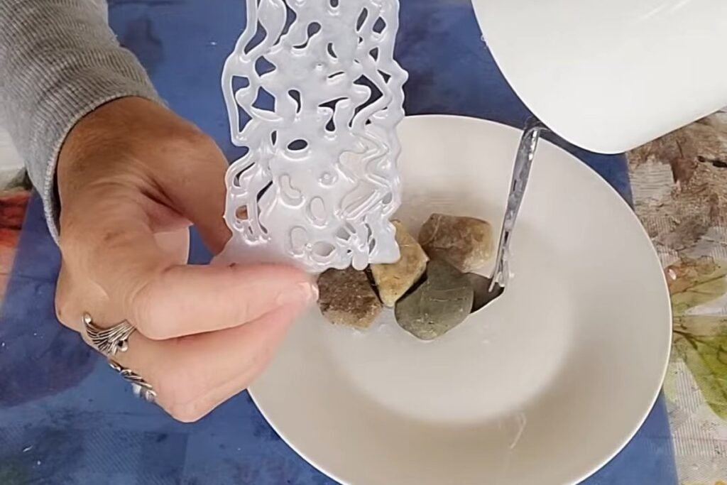 A hand holding a long lacy ribbon of clear hot glue over a saucer with pebbles