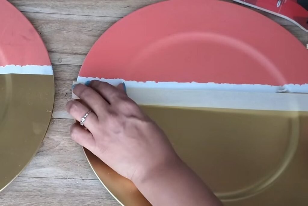 A hand pressing masking tape in a straight line across a large plate that is half coral and half gold
