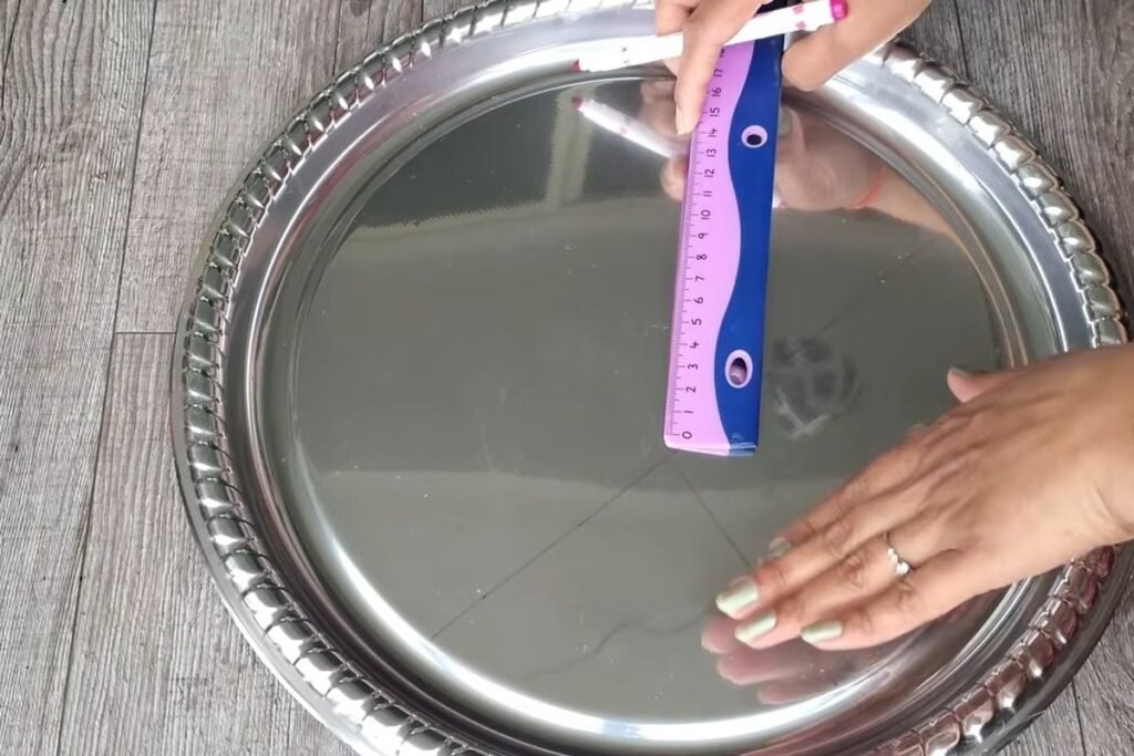 A hand holding a pink ruler across a large shiny metal tray, marking straight lines with a pen