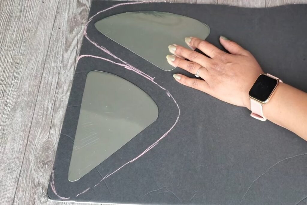 A hand resting on a black foam board where pink outlines have been drawn around two metal mirror pieces