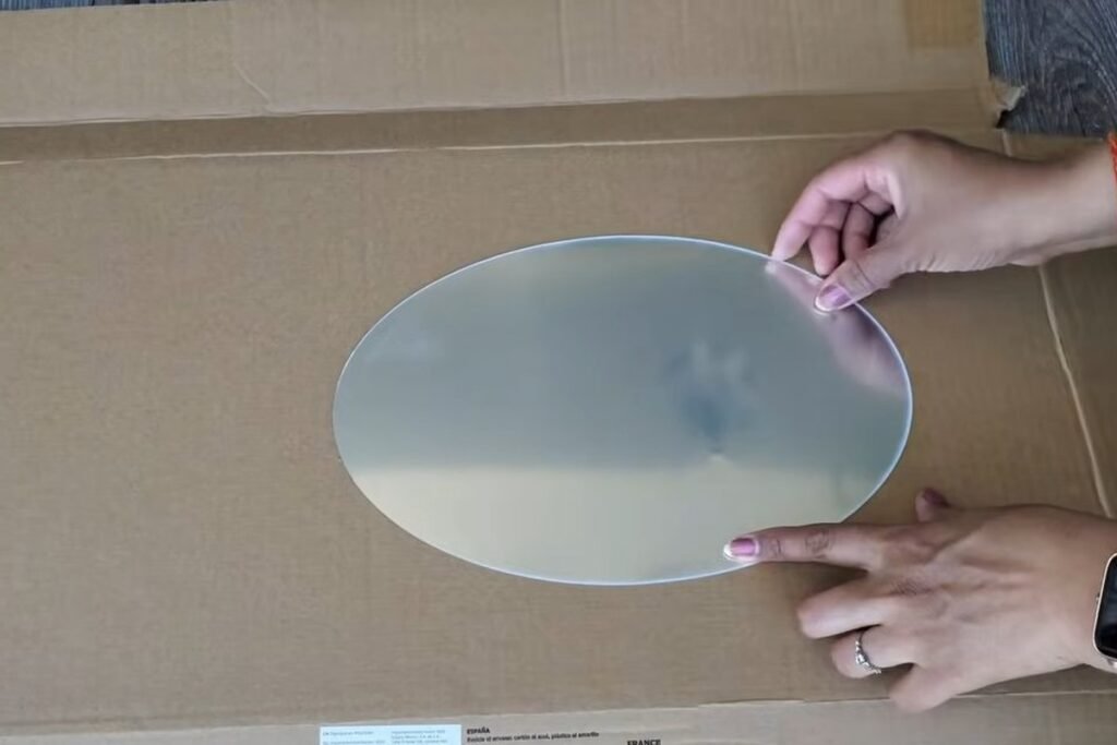 An oval flexible mirror sheet lying on a large piece of cardboard with two hands steadying it