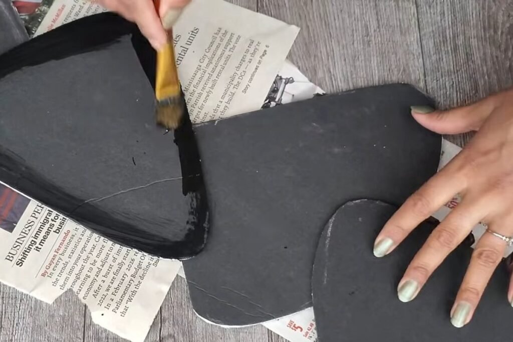 A brush painting black paint along the rim of a black foam shape on a sheet of newspaper