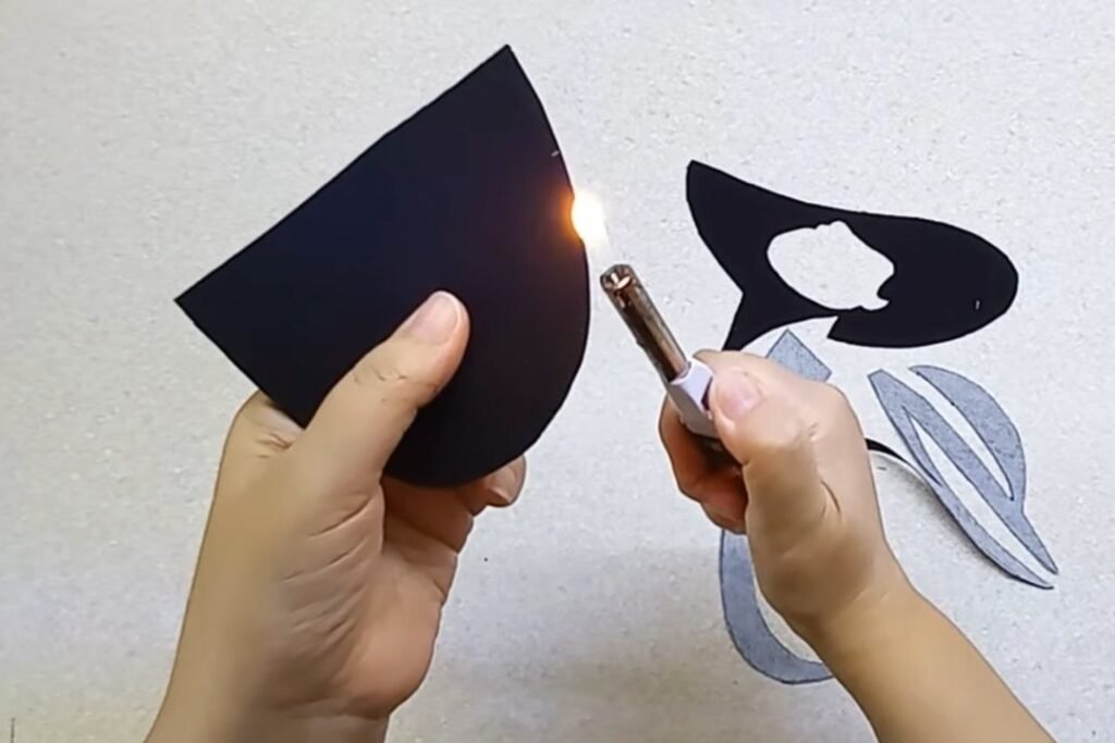 A hand holding a black fabric shape while a lighter flame runs along its edge