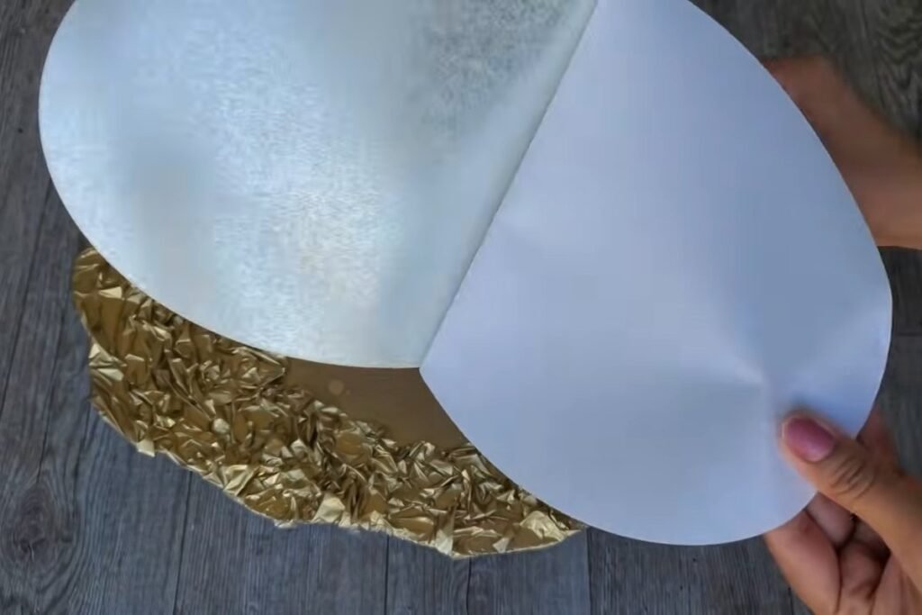 A hand peeling a large white backing sheet away from the back of an oval mirror sheet above the gold frame