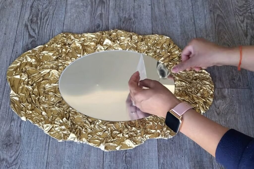 A hand lifting the corner of a thin protective film off the face of a freshly mounted oval mirror in a gold frame