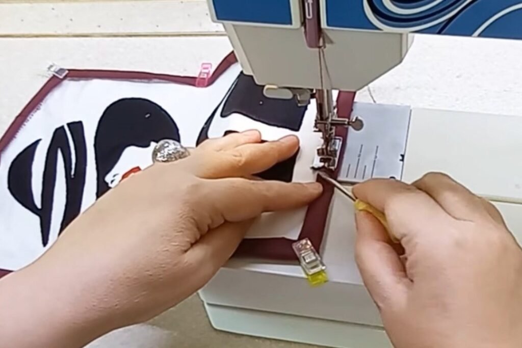 A pink sewing clip holding a maroon border while the sewing machine stitches the edge of a fabric portrait