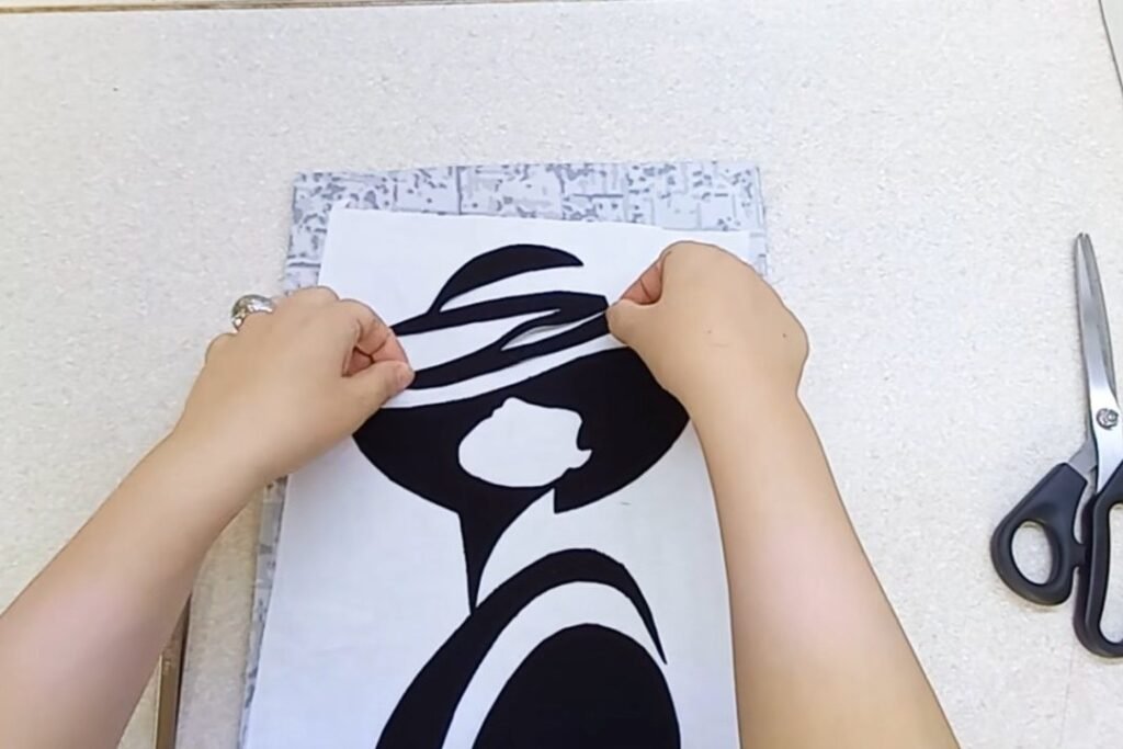Hands placing curved black stripes onto the hat of a fabric portrait with scissors beside it