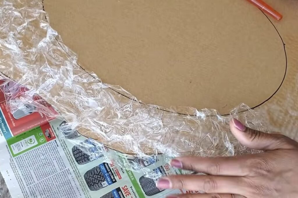 Hands pressing crumpled clear plastic wrap onto a glued cardboard border so it forms deep wrinkles