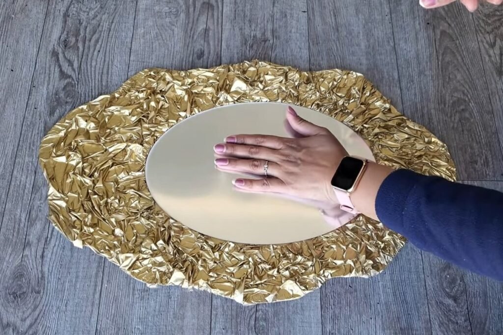A hand pressing a pale oval mirror sheet into the centre of a gold crinkled frame