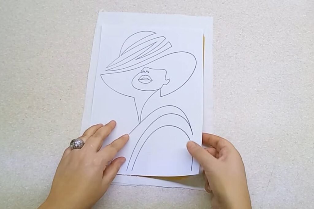 A printed line drawing of a woman in a hat held over white fabric with a yellow sheet underneath