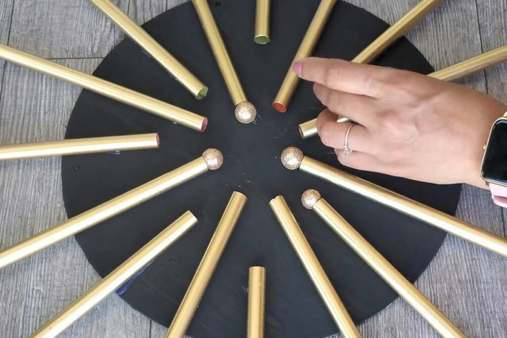 A hand pressing a small gold glitter ball onto the inner end of a gold straw at the centre of the circle