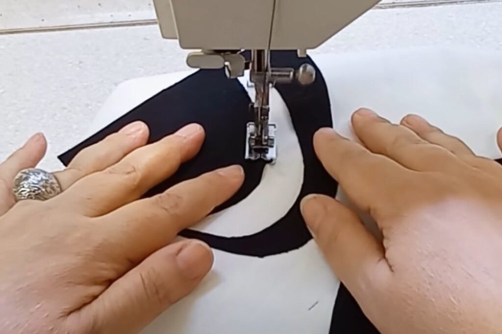 A sewing machine presser foot stitching along the edge of a black fabric shape on white fabric