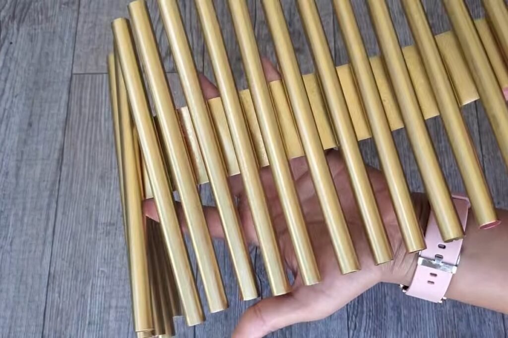 A flat mat of gold painted straws held up by a hand over a wooden floor