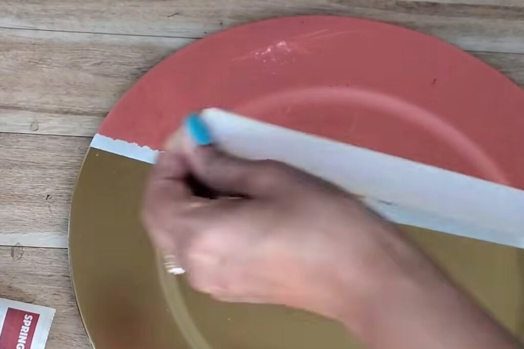 A hand laying a wide strip of masking tape across the gold half of a plate that has a coral top