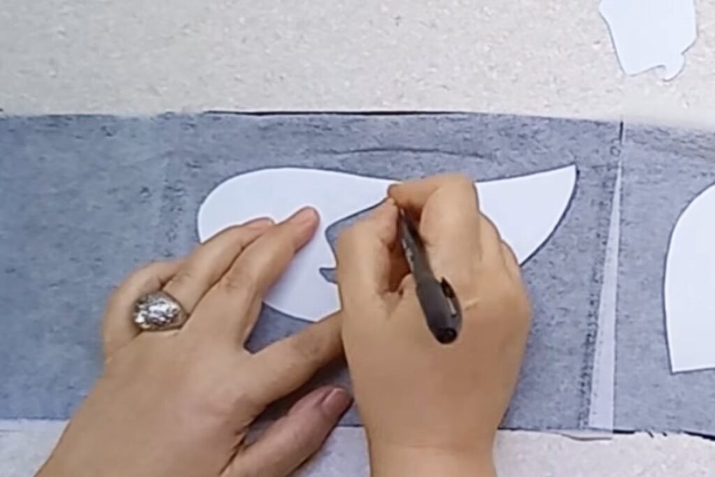 A hand tracing a white paper hat shape onto grey interfacing with a pen