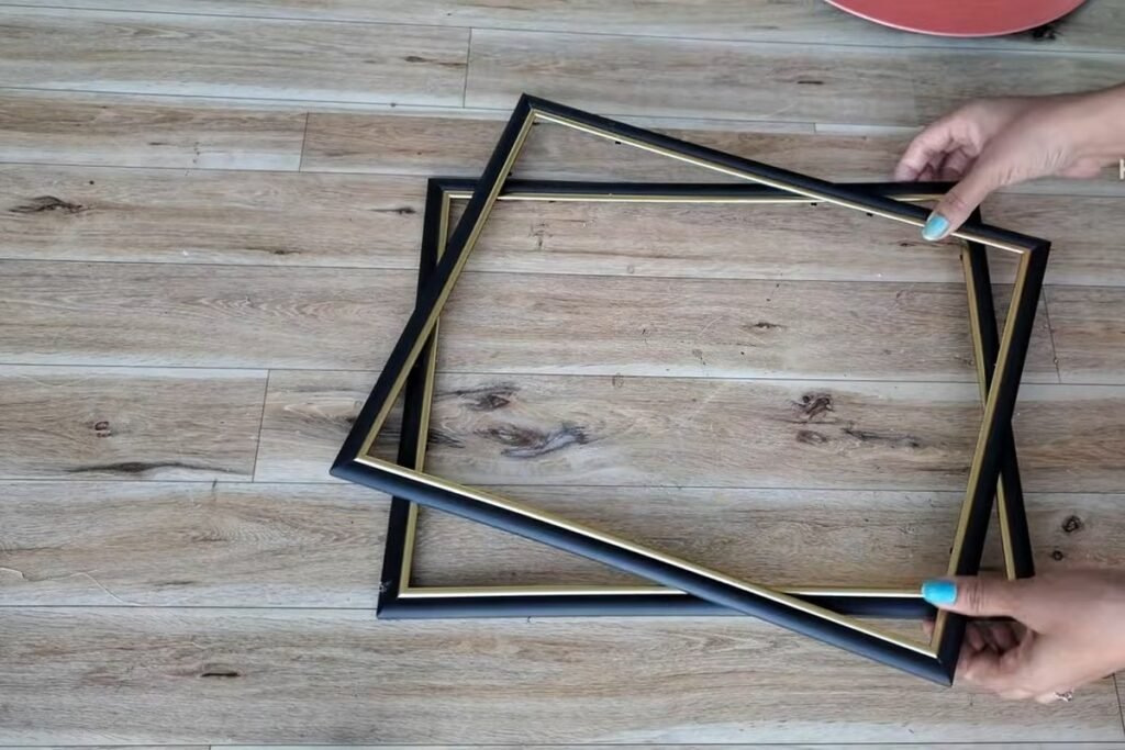 Two empty black picture frames with thin gold inner trim lying on a wooden floor with a hand holding one
