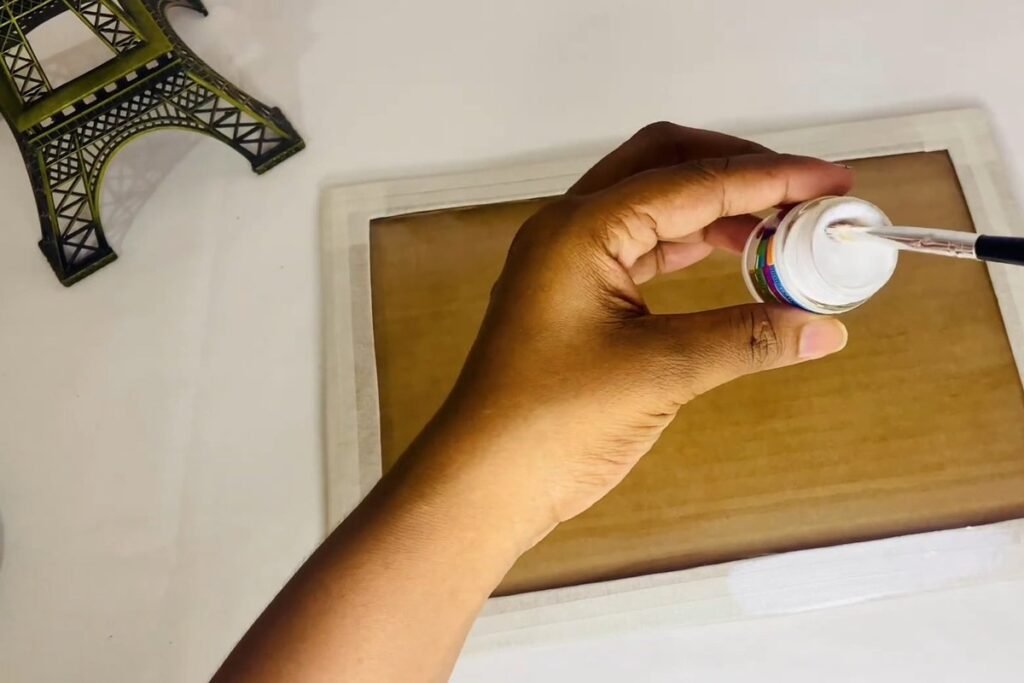 Hand dipping a small brush into a bottle of white fabric paint over a taped frame