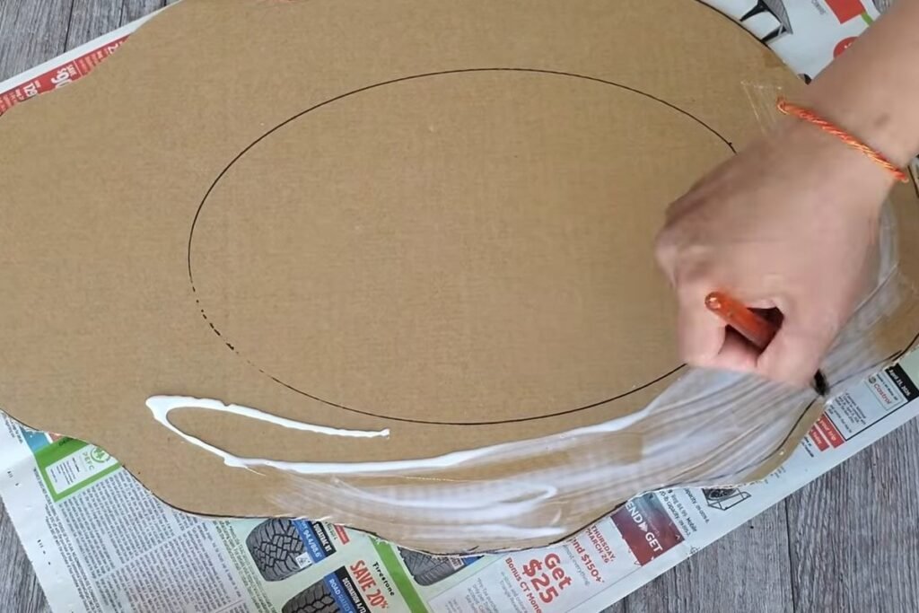 A hand spreading white glue over the border of a cut wavy cardboard shape that rests on newspaper